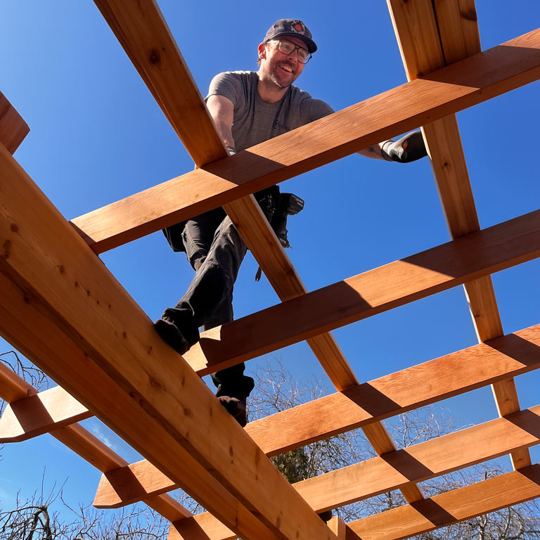A low-angle shot of a smiling man wearing glasses and a baseball cap, standing atop the wooden beams of a pergola under construction. He looks down at the camera against a brilliant, clear blue sky.