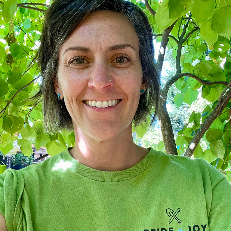 Smiling woman with short hair wearing a green t-shirt, standing outdoors among leafy trees.