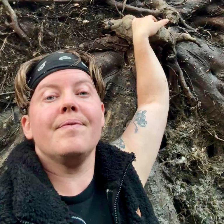 A person with short hair and a black bandana looks at the camera while standing in a forest. They are wearing a black fleece vest, and their arm is raised to touch the dark, tangled roots of an uprooted tree that fills the background.